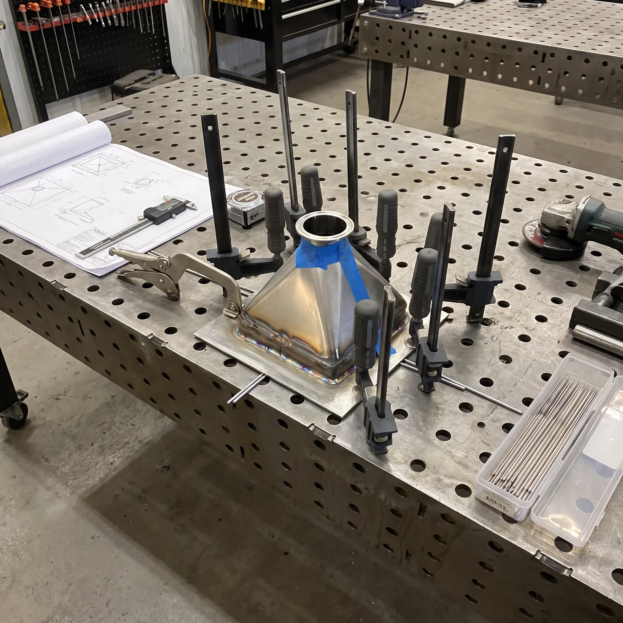 Custom welded tooling fixture beside technical drawings in a fabrication workspace