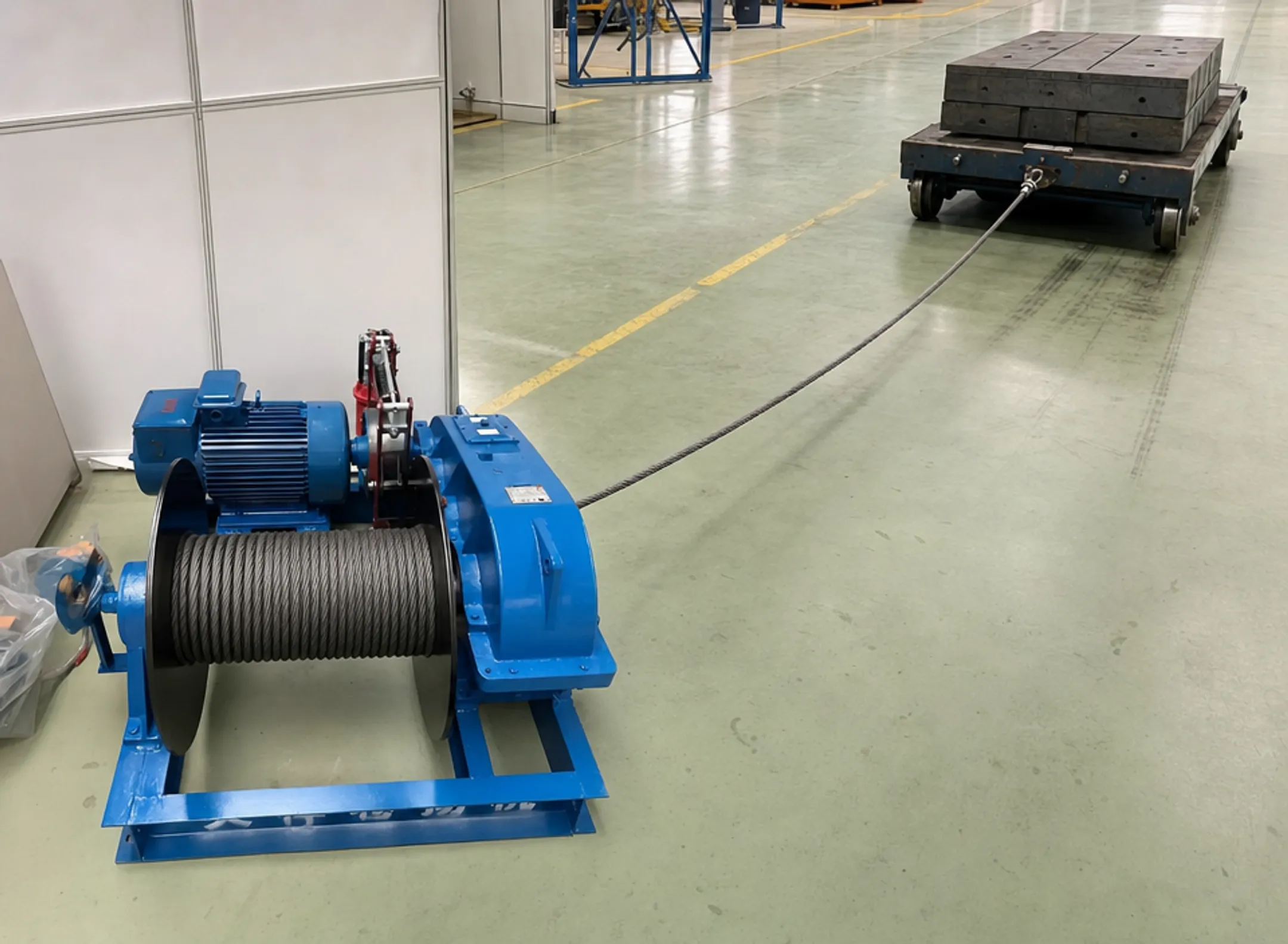 Industrial wall-mounted winch pulling heavy steel cart in facility