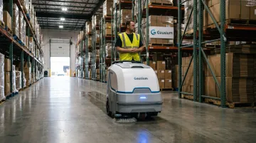 Warehouse worker monitoring autonomous floor cleaning robot operating during shift lull