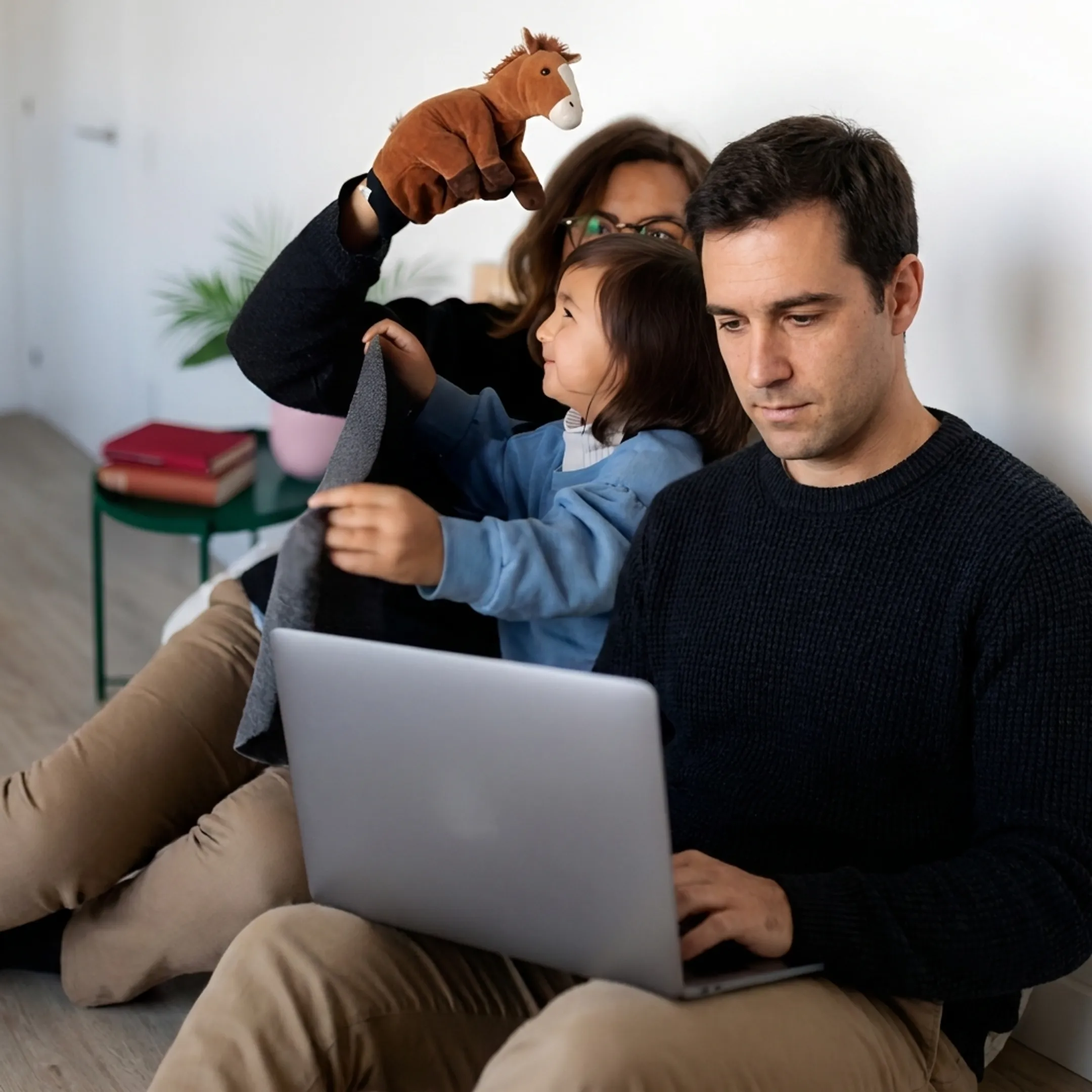 Working parent smiling while working productively at home as a caregiver attends to a young child nearby.