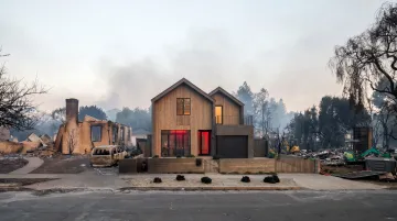 Steel frame home standing intact in wildfire-burned neighborhood with destroyed wood structures nearby