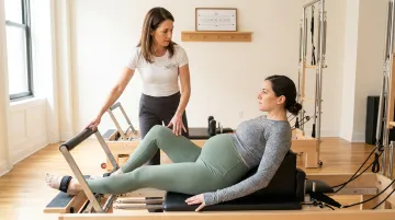Pregnant woman performing modified Pilates Reformer exercise with instructor guidance