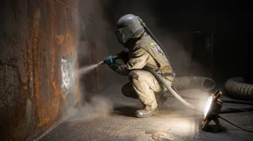 Shipyard abrasive blaster in full PPE operating nozzle inside confined vessel space